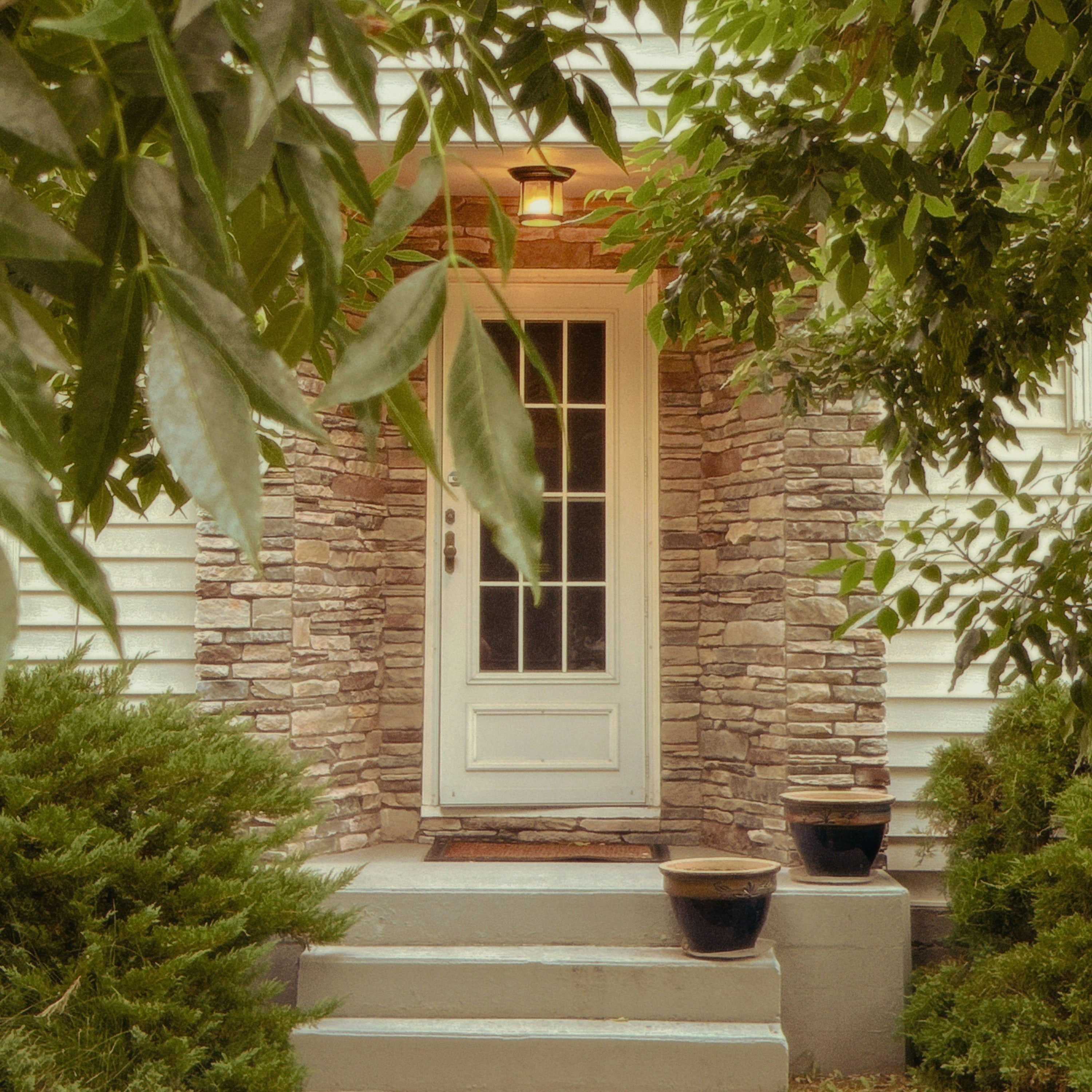 A cream front door with a glowing porch lamp, set in a stone entry and framed by green leaves